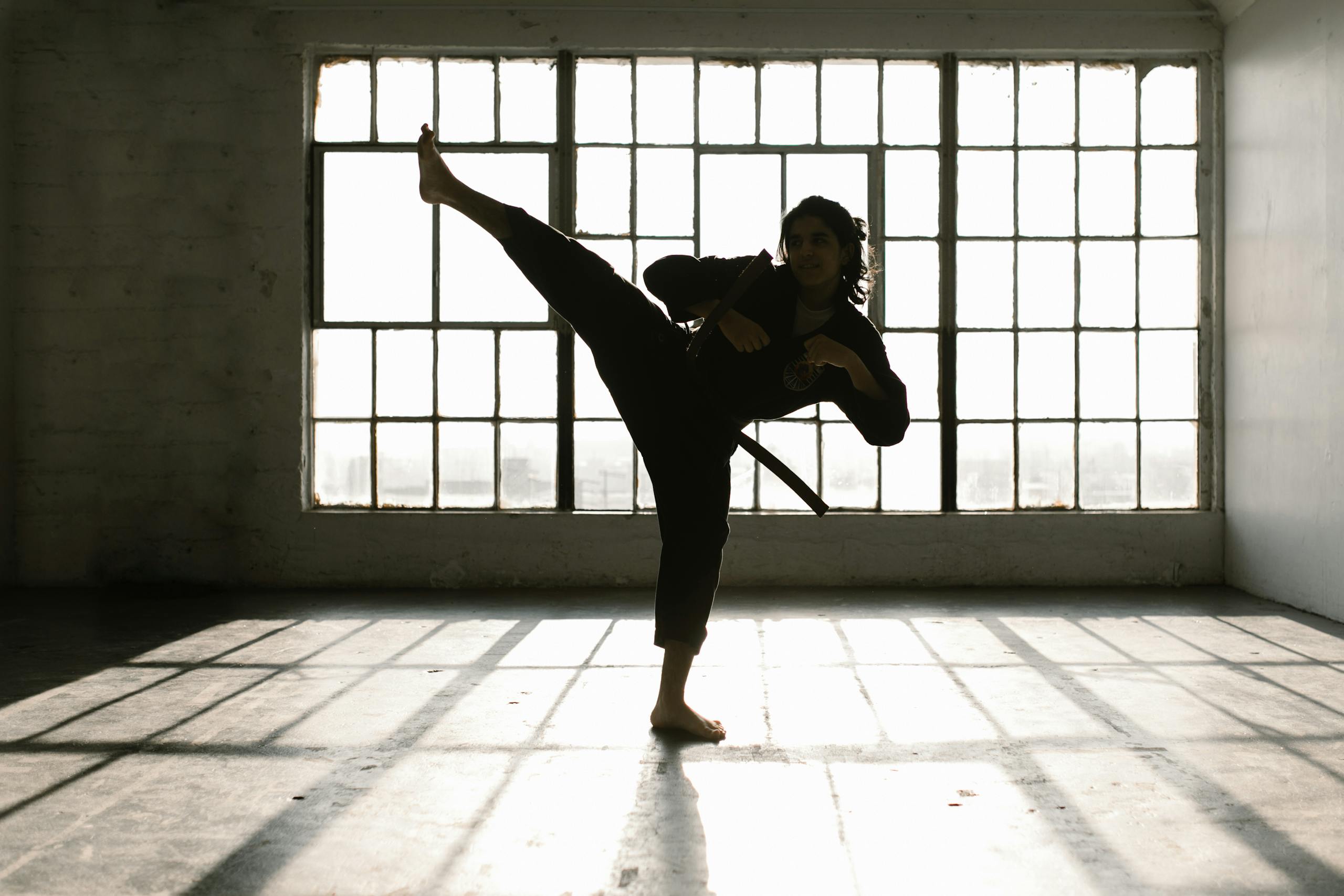 Martial artist performing a high kick in a sunlit room creating dramatic shadows.