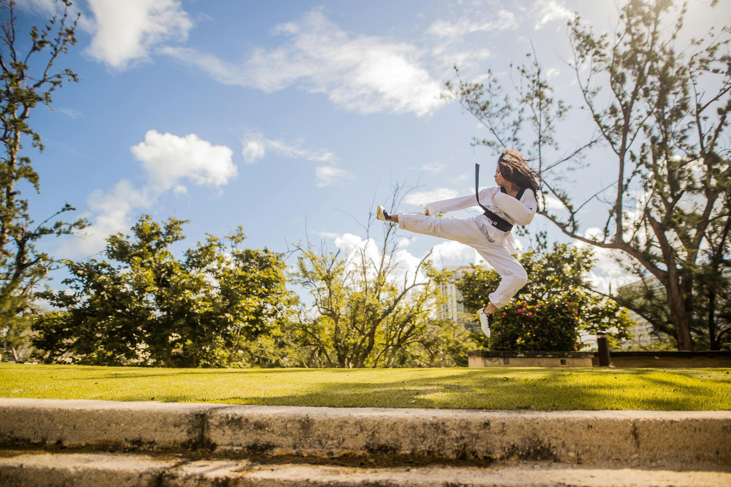 Young woman performs a martial arts kick in a sunny park, showcasing athleticism and focus.